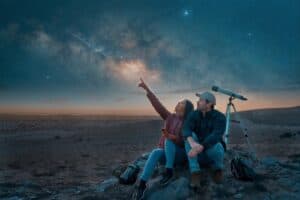 A couple sitting on rocky terrain under a stunning night sky, gazing at a meteor shower with a telescope nearby, symbolising the beauty of stargazing in Ireland.