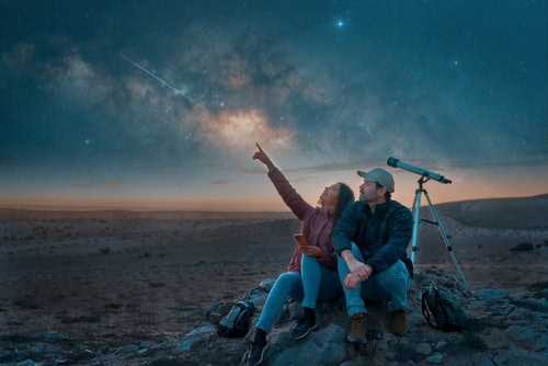 A couple sitting on rocky terrain under a stunning night sky, gazing at a meteor shower with a telescope nearby, symbolising the beauty of stargazing in Ireland.