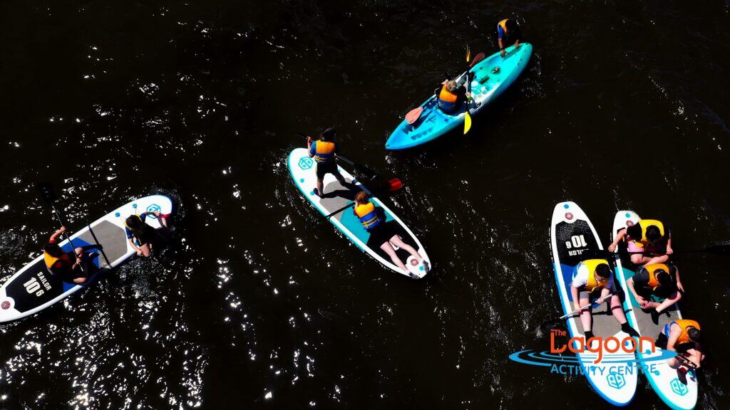 Aerial view of teenagers paddleboarding and kayaking on calm dark water at The Lagoon Activity Centre in West Cork, all wearing life jackets and enjoying a group water activity.