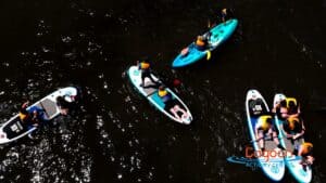 Aerial view of teenagers paddleboarding and kayaking on calm dark water at The Lagoon Activity Centre in West Cork, all wearing life jackets and enjoying a group water activity.